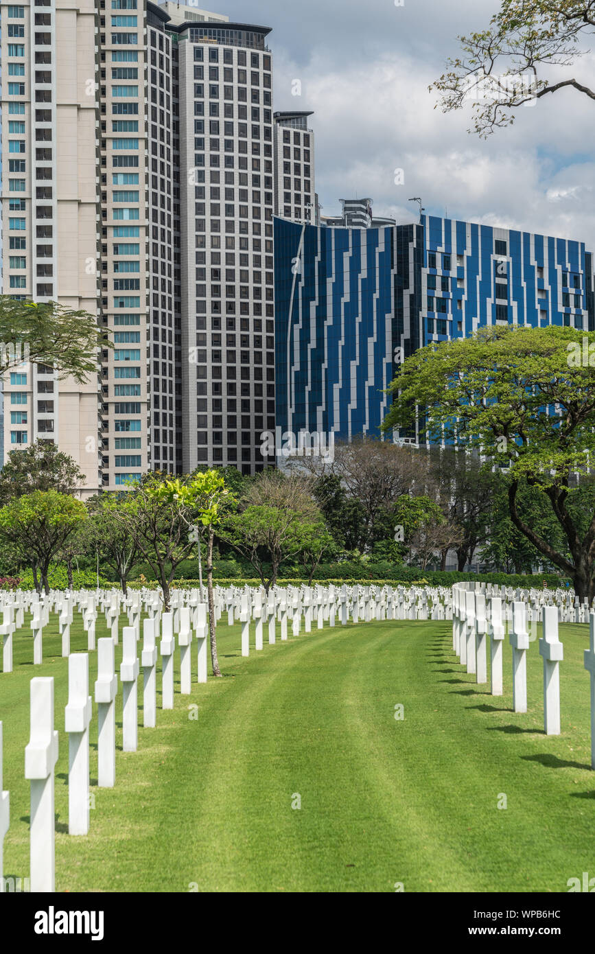 Manila, Filippine - Marzo 5, 2019: Cimitero Americano e il Memorial Park. Grattacieli torreggianti su campo verde riempito con filari di croci bianche. Tre Foto Stock