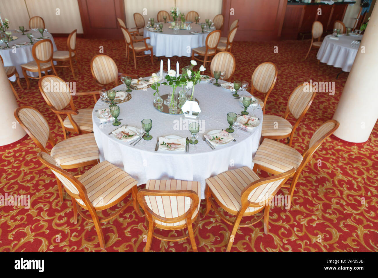 Round tavoli da pranzo, coperto con un panno bianco, serviti con bicchieri di verde e ornate di fiori, stare in piedi su un tappeto di colore. Foto Stock