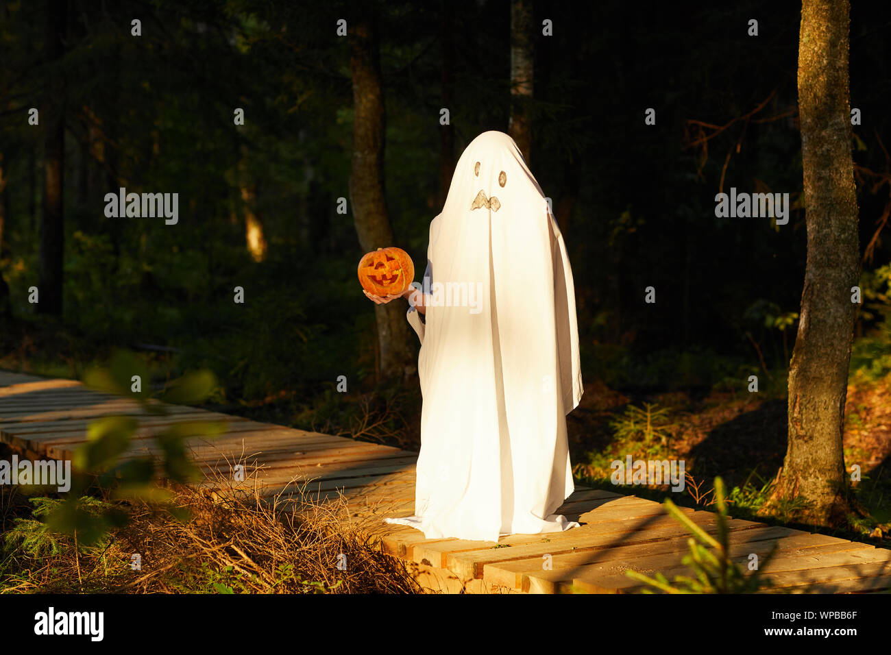 A piena lunghezza Ritratto di spooky bambino vestito come fantasma tenendo la zucca in posa all'aperto di Halloween, spazio di copia Foto Stock