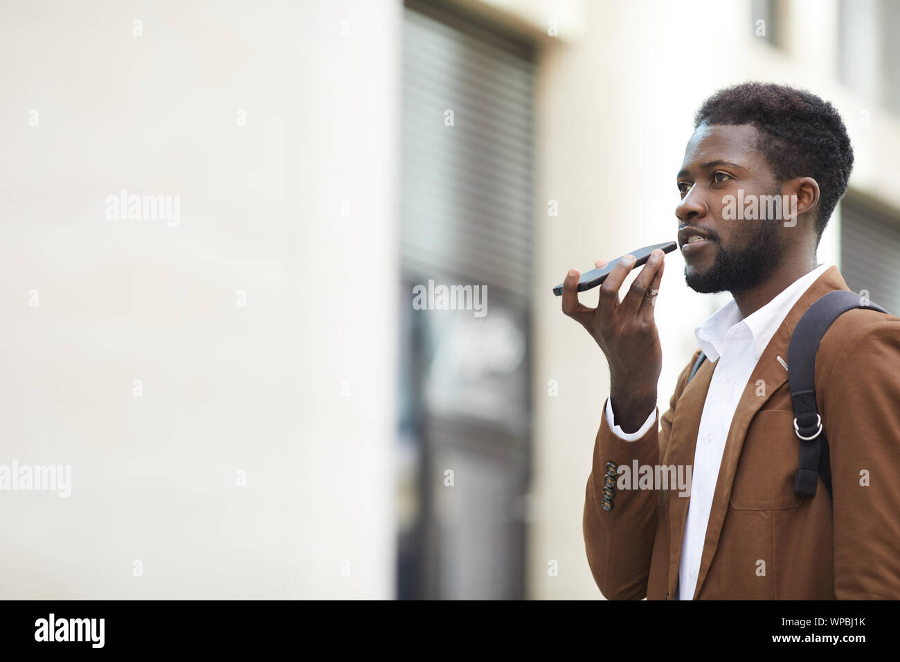 Vista laterale verticale del contemporaneo afro-uomo la registrazione di messaggi vocali tramite smartphone mentre si cammina in una strada di città, spazio di copia Foto Stock