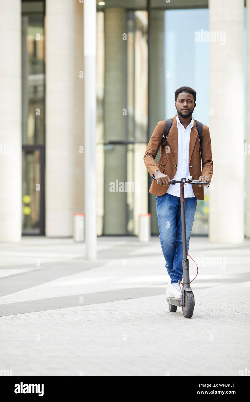 A piena lunghezza Ritratto di contemporaneo afro-uomo che cavalca scooter elettrico e guardando la telecamera durante gli spostamenti nelle strade cittadine, spazio di copia Foto Stock