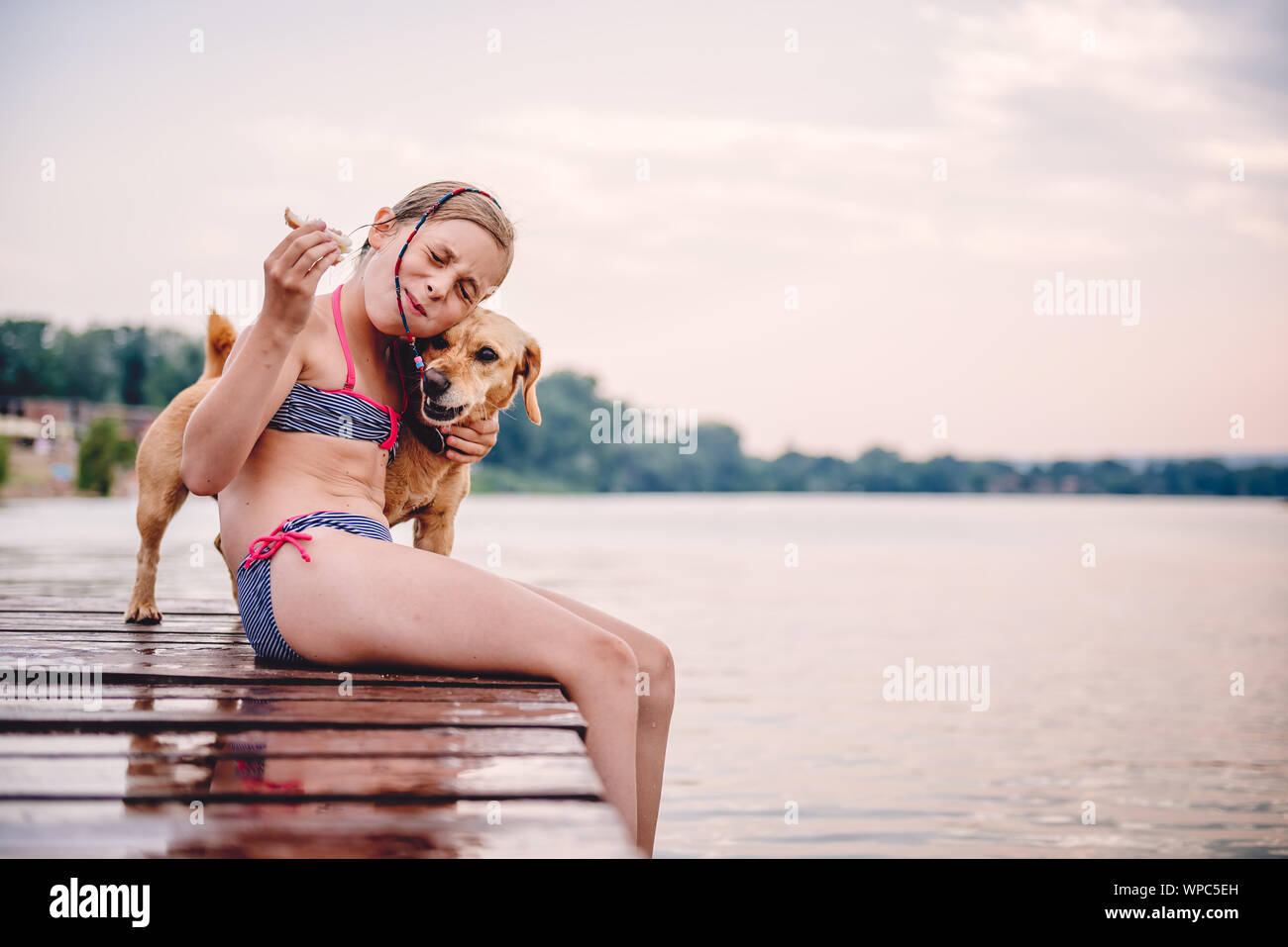 Bambina che abbraccia il suo cane sul dock e mangiare panino Foto Stock Bambina che abbraccia il suo cane sul dock e mangiare panino Foto Stock
