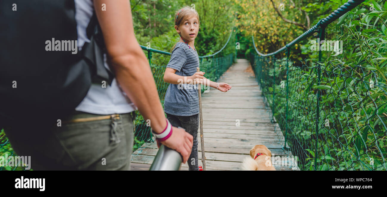 Madre e figlia camminare su legno ponte di sospensione con un piccolo cane giallo nella foresta Foto Stock