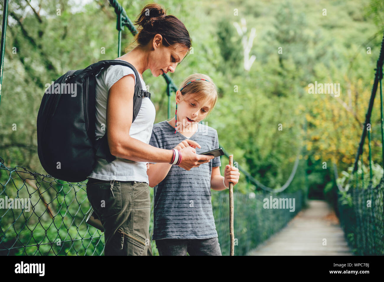 Madre e figlia in piedi sul legno ponte di sospensione nella foresta e utilizzo di smart phone per navigare Foto Stock