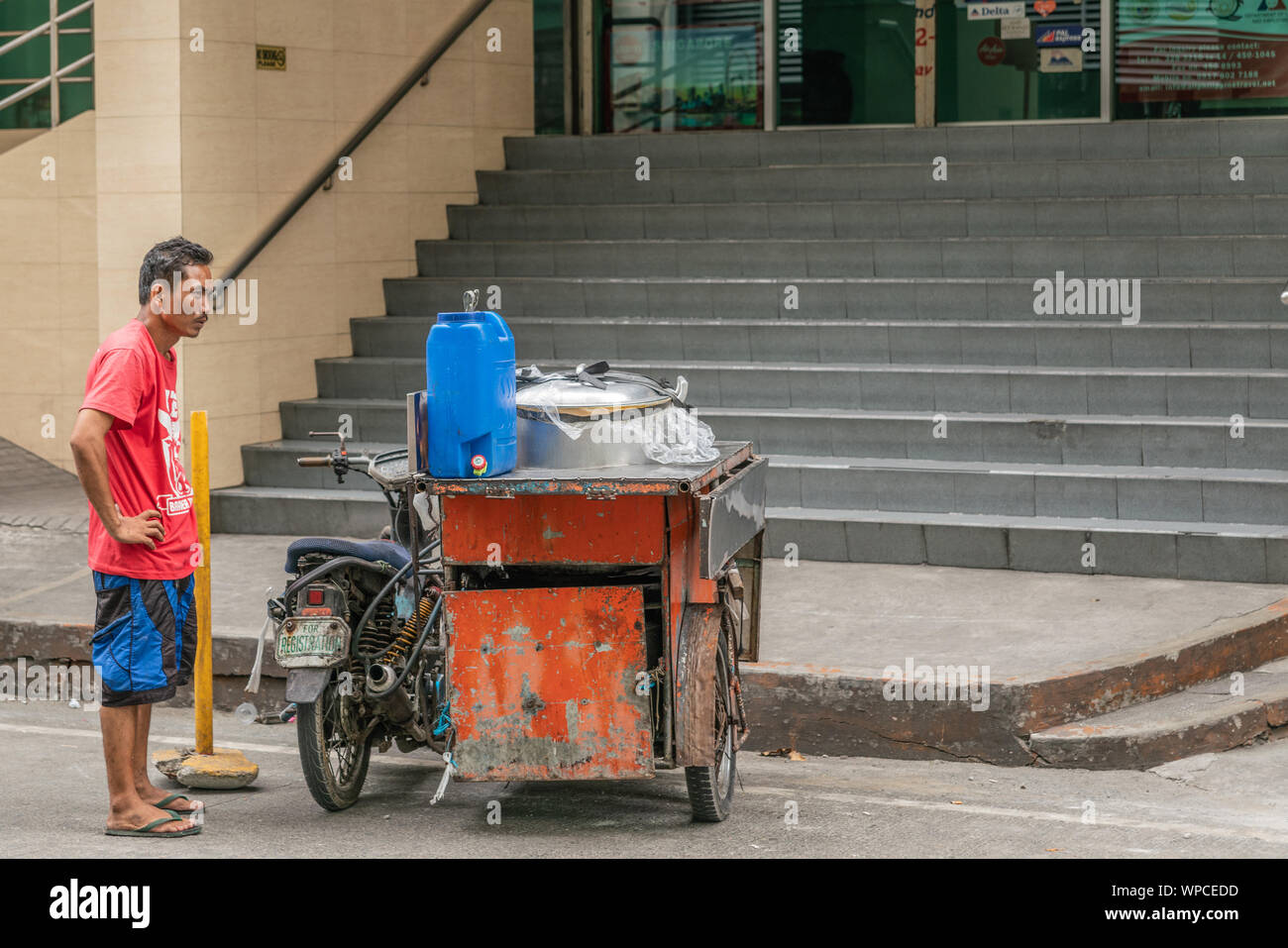 Manila, Filippine - Marzo 5, 2019: ambulatoriamente street food fornitore con red arrugginita triciclo attende i clienti verso il basso il grigio a fasi di costruzione di ufficio o Foto Stock