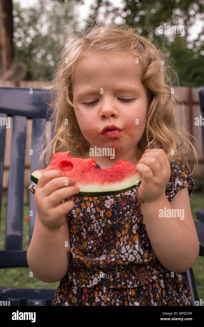 Incantevole piccola ragazza con i capelli ricci, mangiare succoso, delizioso e fresco cocomero, con espressione divertente Foto Stock