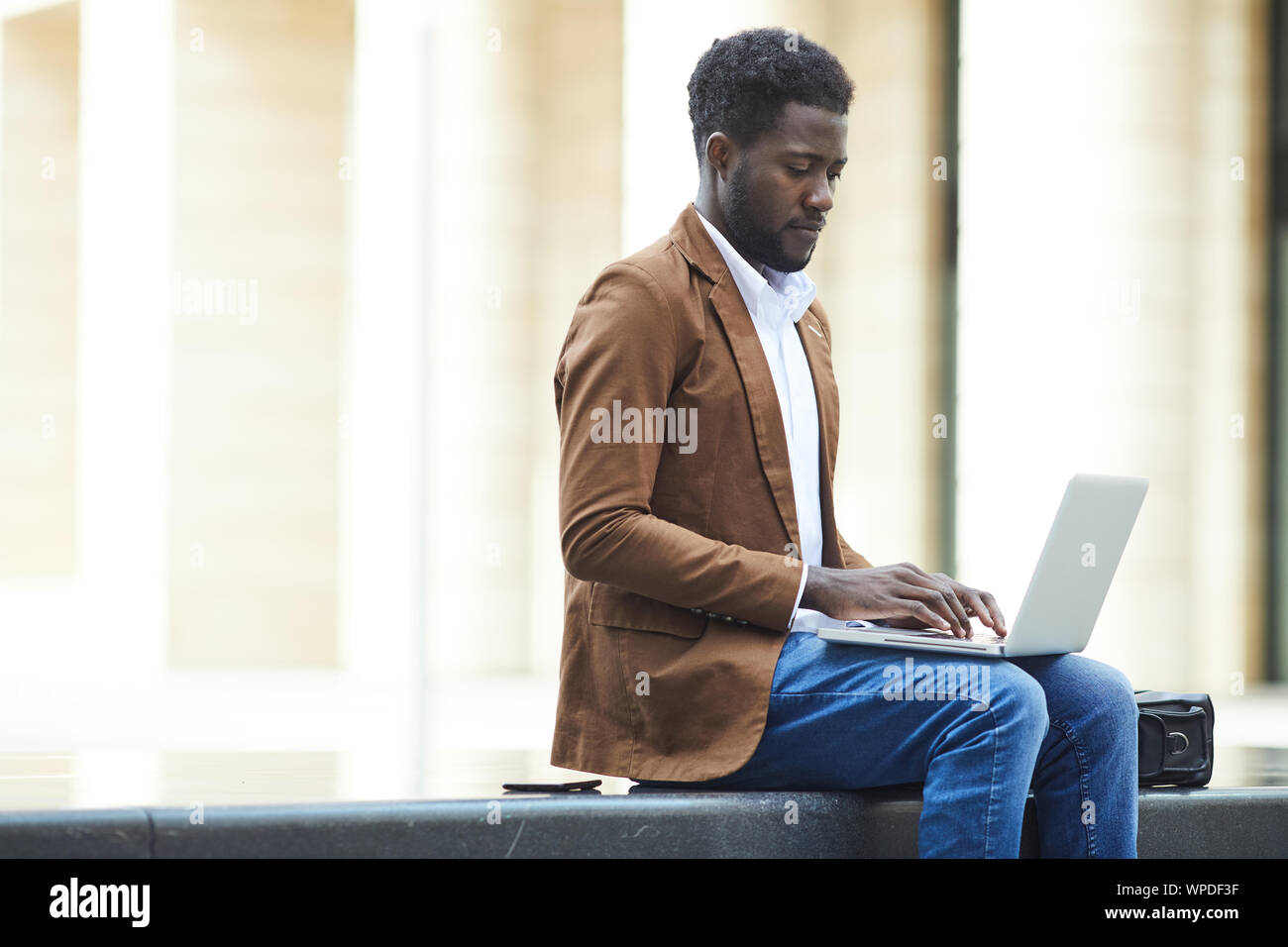 Vista laterale Ritratto di giovane uomo afro-americano utilizzando laptop all'aperto mentre si lavora sul progetto di business nel contesto urbano, spazio di copia Foto Stock