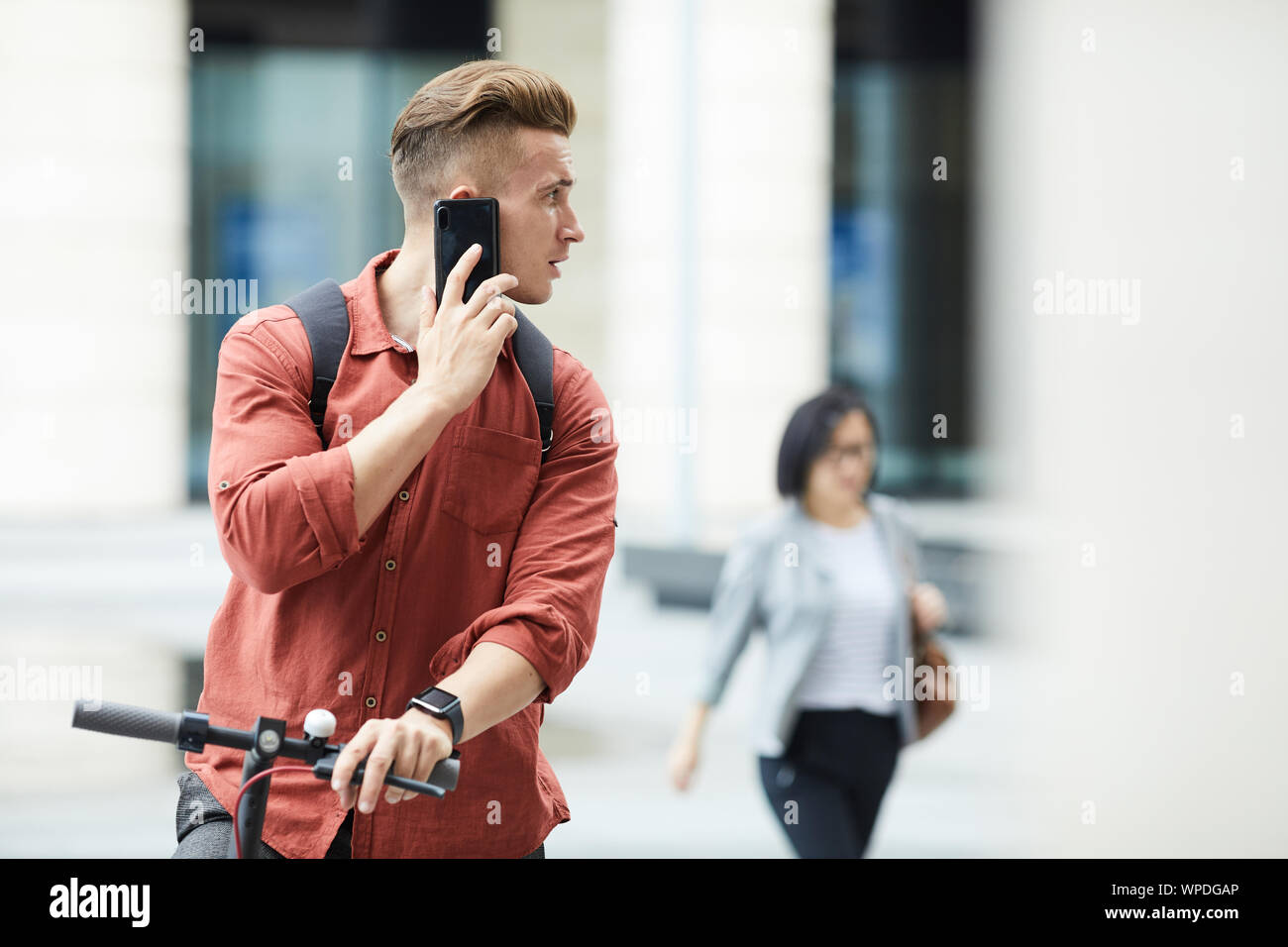 Vita ritratto di bel giovane che parla tramite smartphone mentre cavalcate scooter elettrico nel contesto urbano, spazio di copia Foto Stock