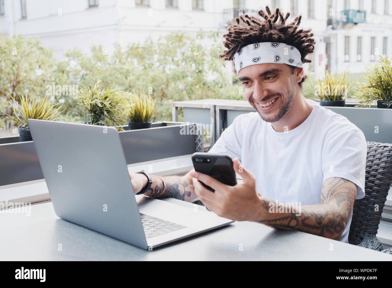 Sorridente giovane maschio freelancer digitando su computer portatile durante la lettura del messaggio sul reddito sullo smartphone, positivo hipster guy verifica delle notifiche su mobi Foto Stock