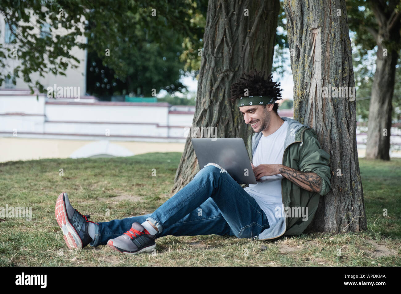 Bel giovane studente internazionale seduti su erba nel parco pubblico, la digitazione di un messaggio di testo sulla tastiera del laptop moderno computer, chiacchierando in ne sociale Foto Stock
