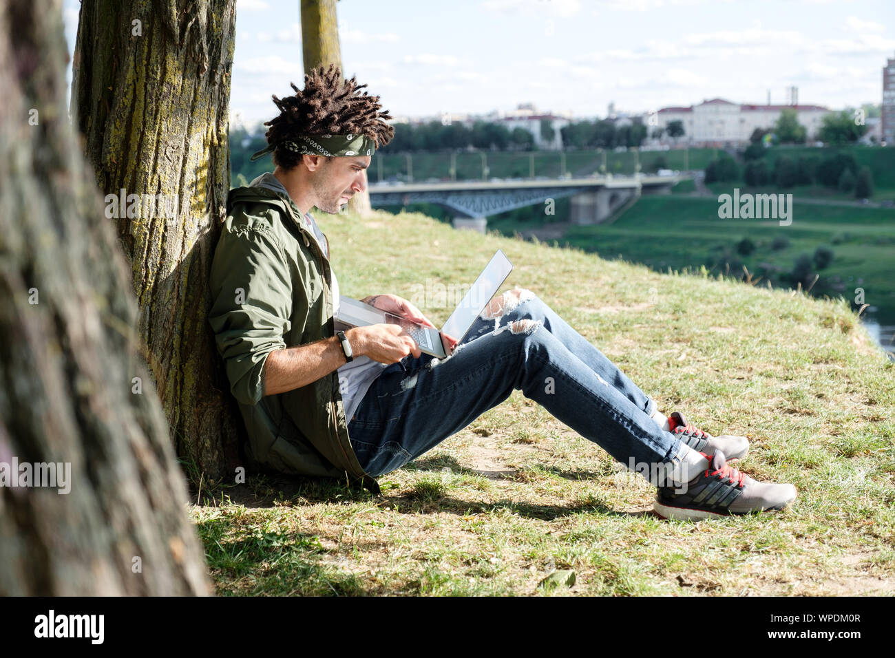 Giovane studente millenario con dreadlocks seduto su erba e utilizzando computer portatile laptop all'aperto nel parco pubblico. Libero professionista utilizzando notebook per dis Foto Stock