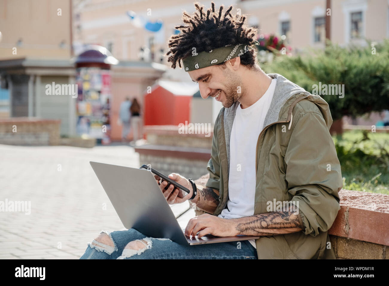 Vista laterale colpo di un uomo bello utilizza lo smartphone mentre si lavora sul computer portatile all'aperto, felice ragazzo con african dreadlocks acconciatura verificare notifica, rea Foto Stock