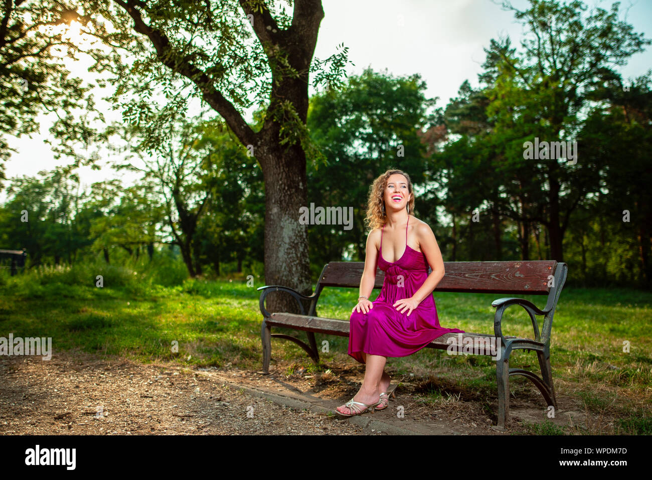 Bellissimi capelli ricci giovane donna indossa elegante vestito viola seduta su una panchina nel mezzo della foresta con raggi di luce del sole durante il tramonto in attesa h Foto Stock