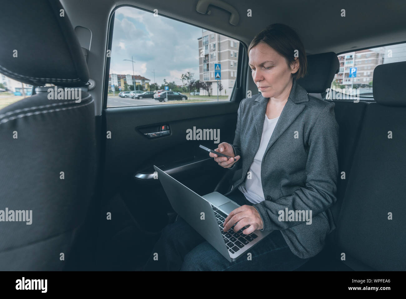 Imprenditrice mediante telefono cellulare e computer portatile in auto tutta la seduta sul sedile posteriore, comunicazione aziendale in movimento Foto Stock