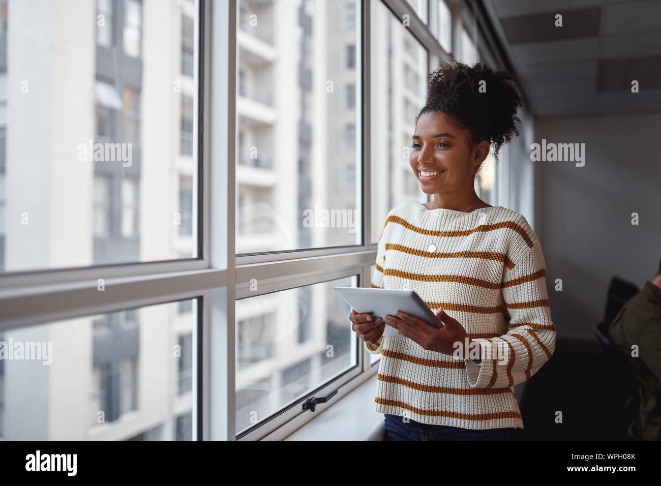 Riflessivo sorridente bella giovane americano africano imprenditrice con tavoletta digitale in piedi alla finestra di ufficio che guarda lontano Foto Stock