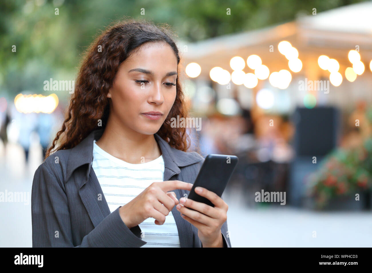 Grave razza mista donna utilizzando smart phone a piedi in strada con le luci della città in background Foto Stock
