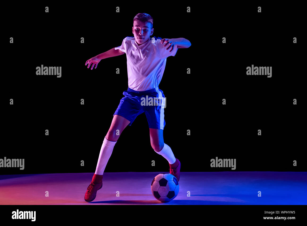 Ragazzo giovane come il calcio o il giocatore di football in sportwear  facendo una finta o un calcio con la palla per un obiettivo di studio scuro  dello sfondo. Montare la riproduzione