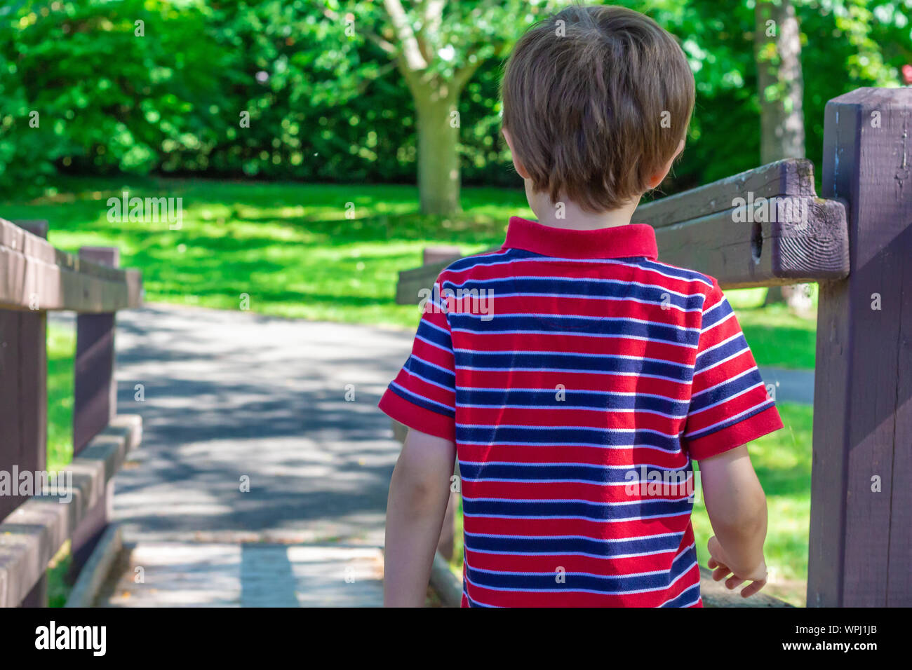 Un giovane ragazzo, circa 5 anni di età, indossa un rosso-blu striped Polo shirt come egli inizia a camminare lungo una rampa di legno in un parco pubblico. Foto Stock