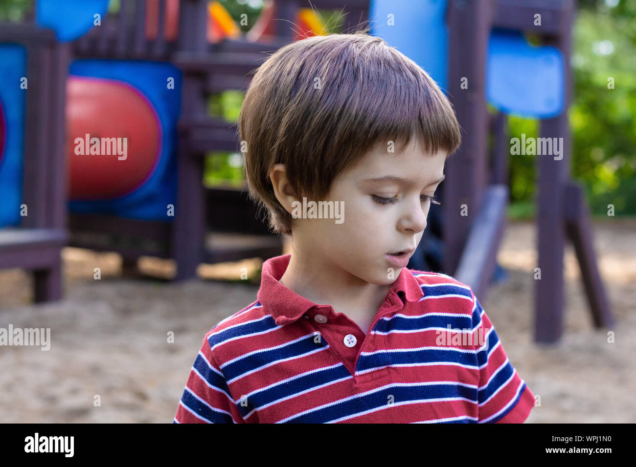 Un ragazzo di circa 5 anni, guarda verso il basso e verso destra come egli è nel profondo del pensiero. Egli si trova di fronte a un parco giochi per bambini palestra nella giungla. Foto Stock