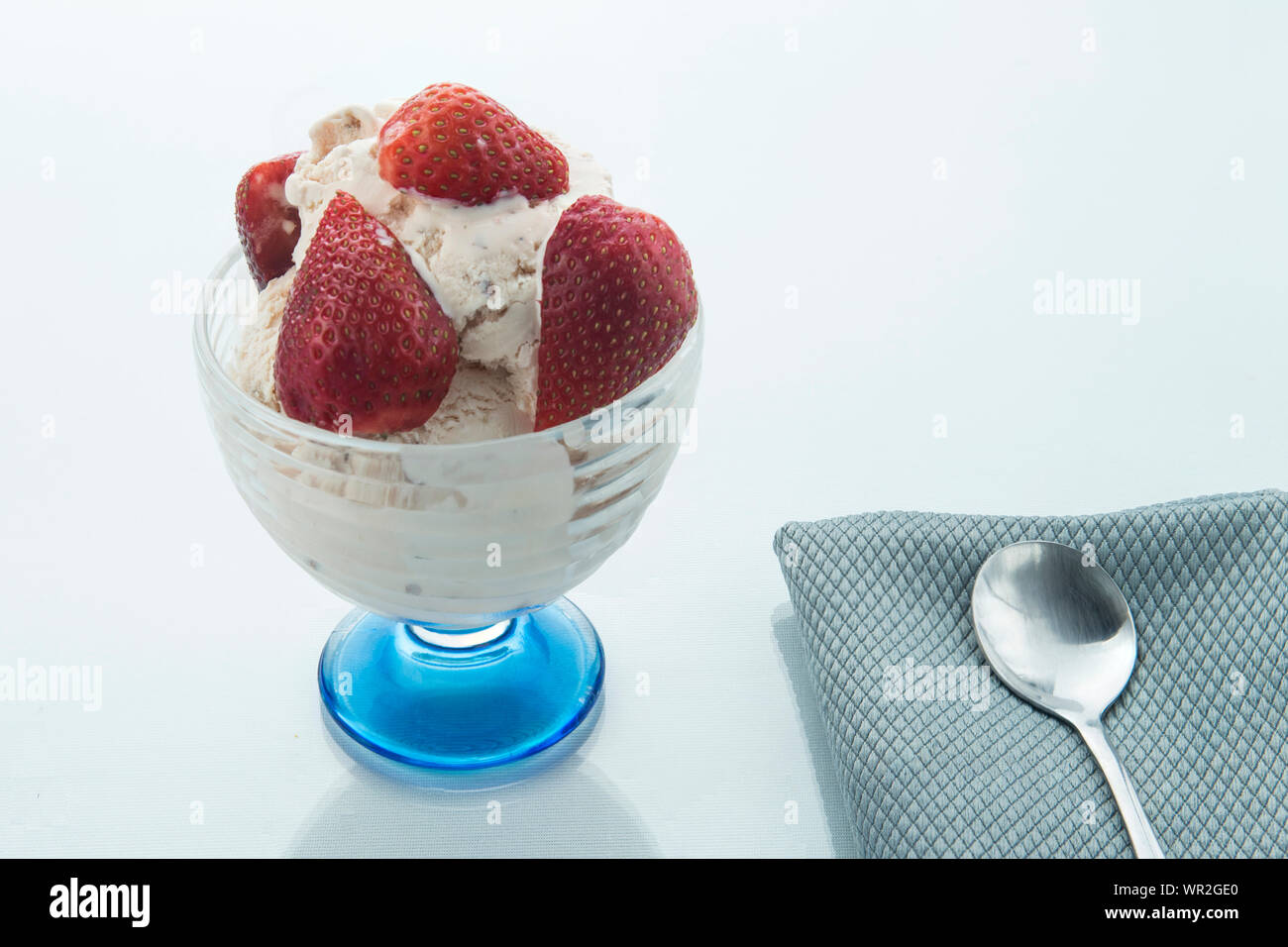 Le fragole in un bicchiere di vetro e più fragole su una piastra blu su bianco e superficie lucida e una forcella sul lato, sfondo bianco. Foto Stock