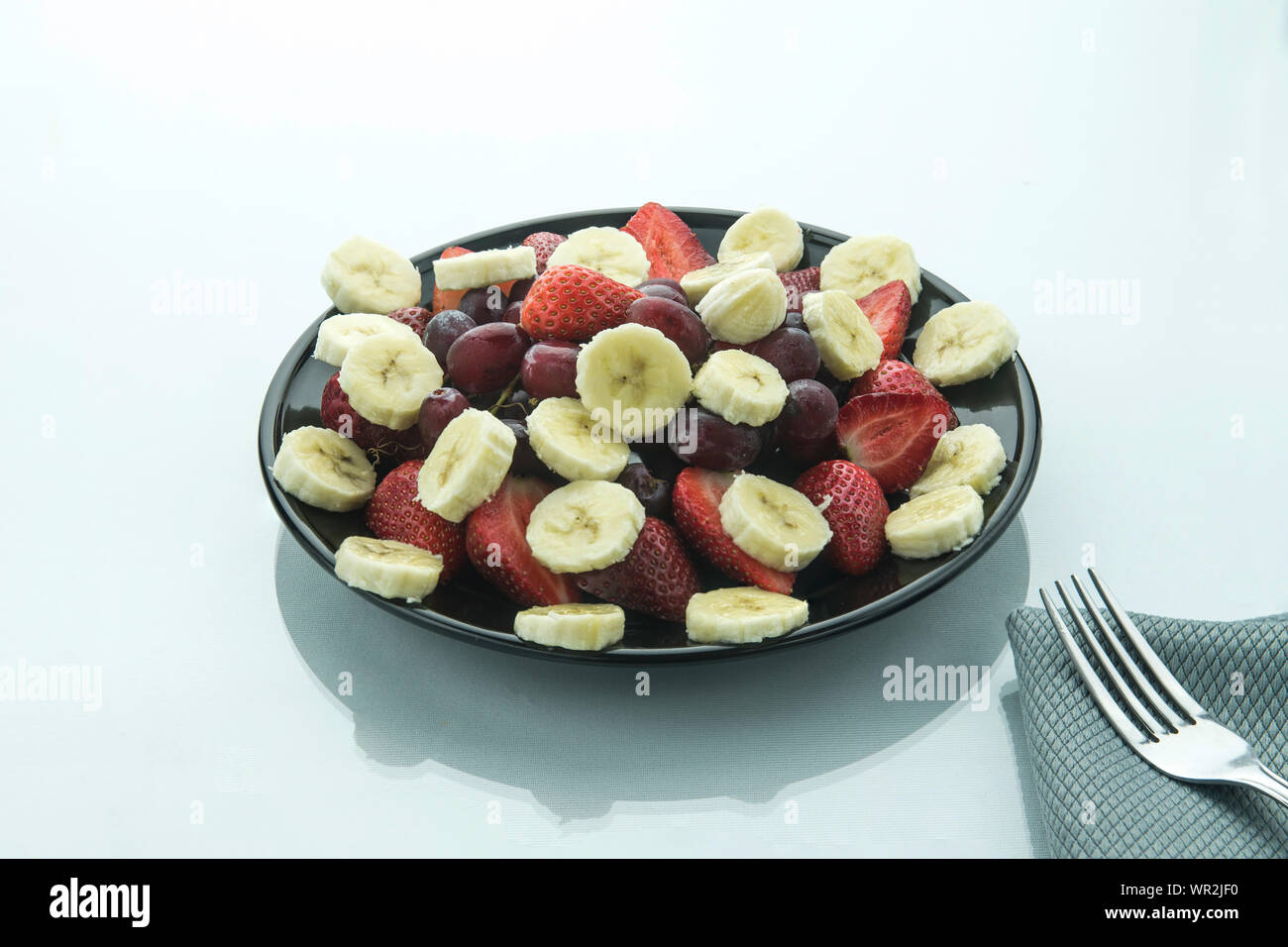 Uva e banana strawbery le fette su un piatto nero su bianco e superficie lucida con una forcella sul lato e sfondo bianco. Foto Stock