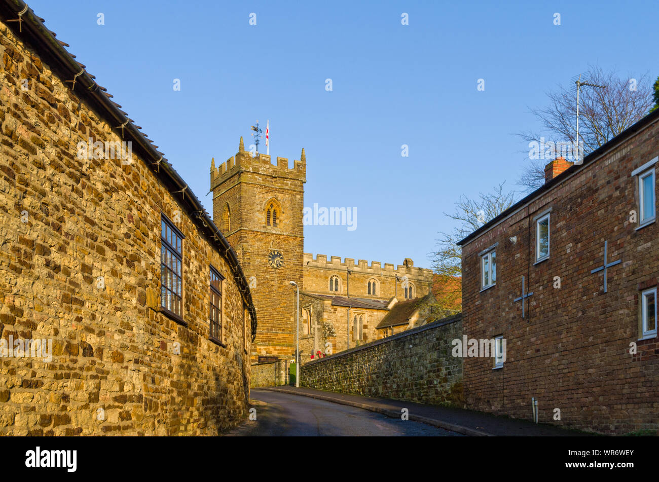 Strada in vista del villaggio di Wootton, Northampton, Regno Unito: ironstone case con la duecentesca chiesa di San Giorgio in distanza. Foto Stock