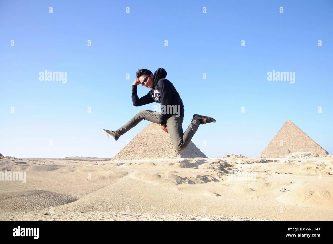 Giovane uomo che indossa Maglia con cappuccio e occhiali da sole a metà aria a piramidi di Giza contro il cielo chiaro Foto Stock