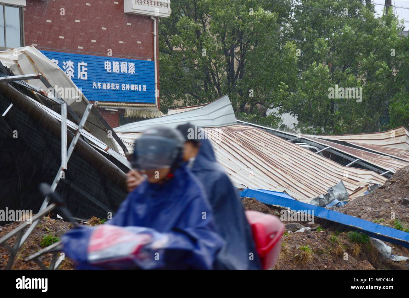 I ciclisti cinesi un giro in scooter in heavy rain passato i detriti di un capannone in acciaio soffiata dal forte vento causato dal tifone Lekima in Zhoushan Foto Stock