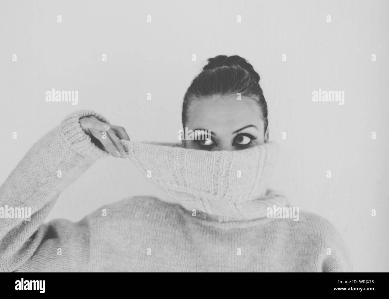 Giovane donna che ricopre la faccia con Turtleneck maglione in piedi contro uno sfondo bianco Foto Stock