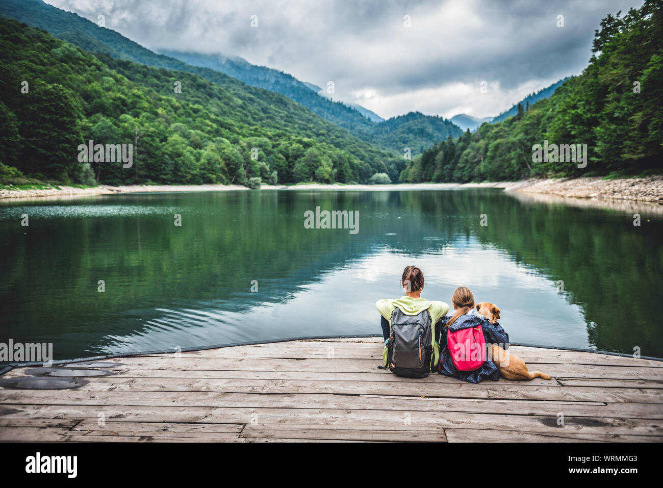 Madre e figlia con un piccolo cane giallo in appoggio su un molo e guardando il lago e le montagne di nebbia Foto Stock Madre e figlia con un piccolo cane giallo in appoggio su un molo e guardando il lago e le montagne di nebbia Foto Stock