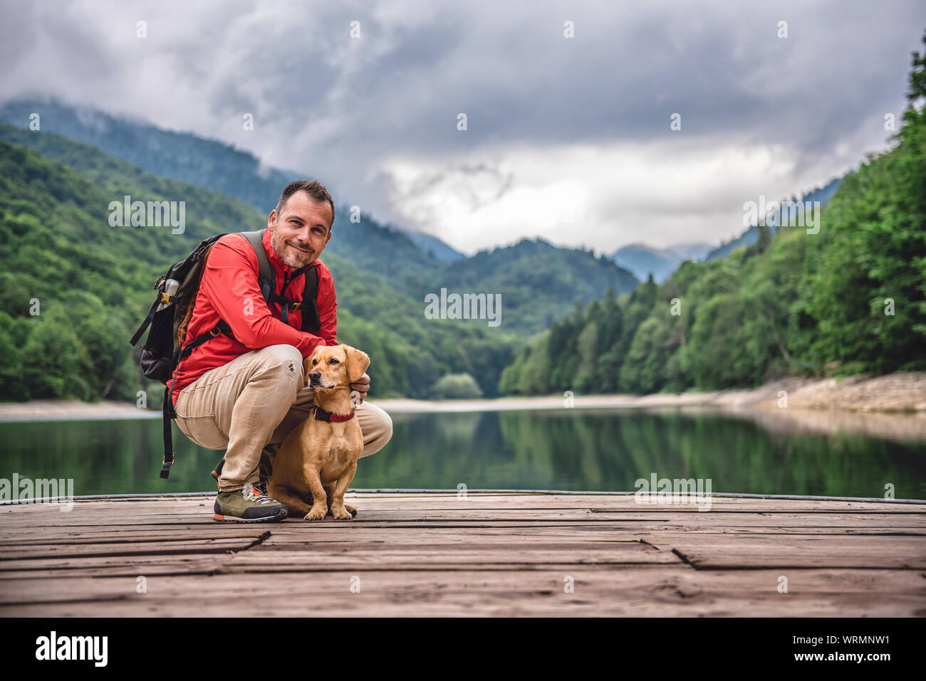 Escursionista indossando giacca rossa e uno zaino con un piccolo cane giallo su un molo in posa dal lago di montagna e la foresta di nebbia Foto Stock Escursionista indossando giacca rossa e uno zaino con un piccolo cane giallo su un molo in posa dal lago di montagna e la foresta di nebbia Foto Stock