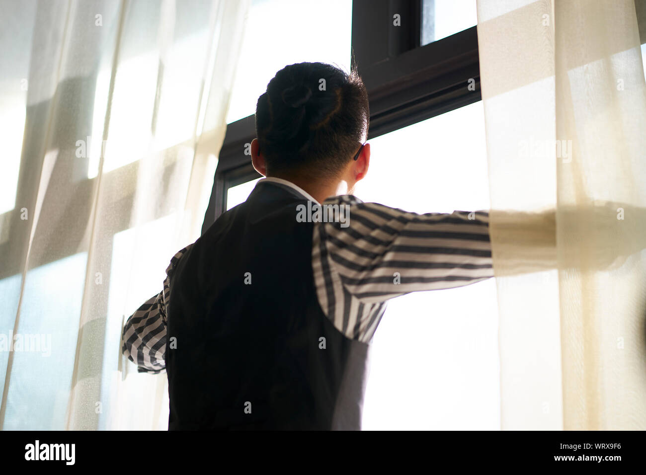 Vista posteriore di un giovane uomo asiatico guardando fuori della finestra Foto Stock
