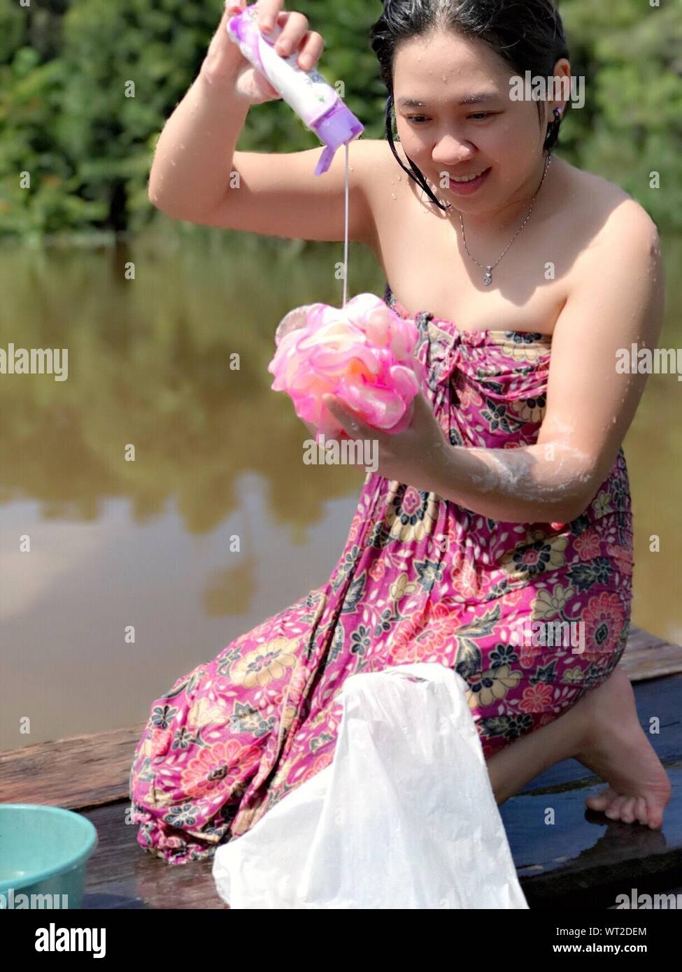 Sorridente giovane donna corpo versatore lozione sulla Luffa mentre inginocchiato al molo sul lago Foto Stock