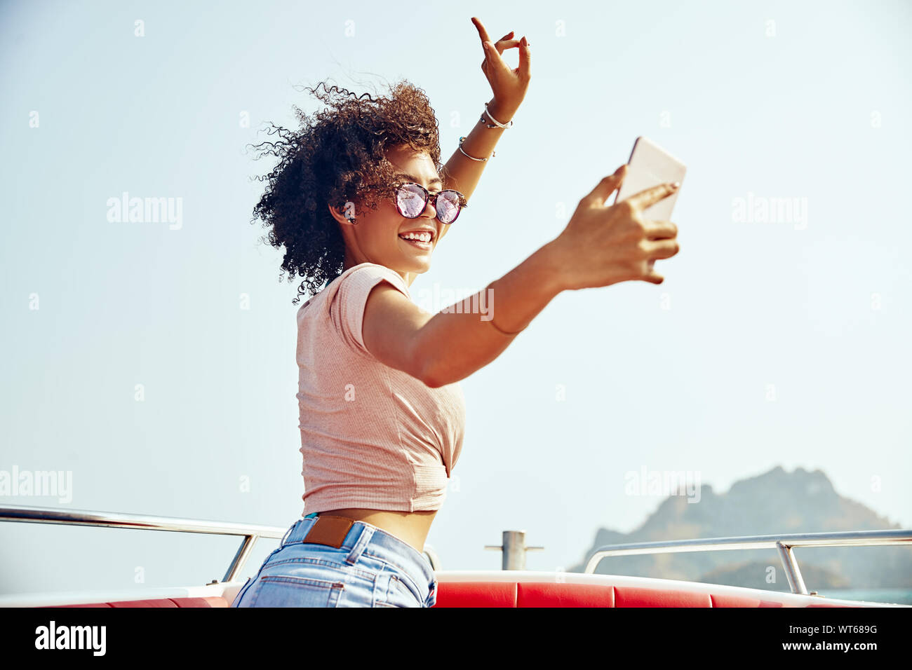 Spensierato giovane donna che indossa gli occhiali da sole in piedi su una barca contro un cielo blu tenendo selfies durante le sue vacanze estive Foto Stock
