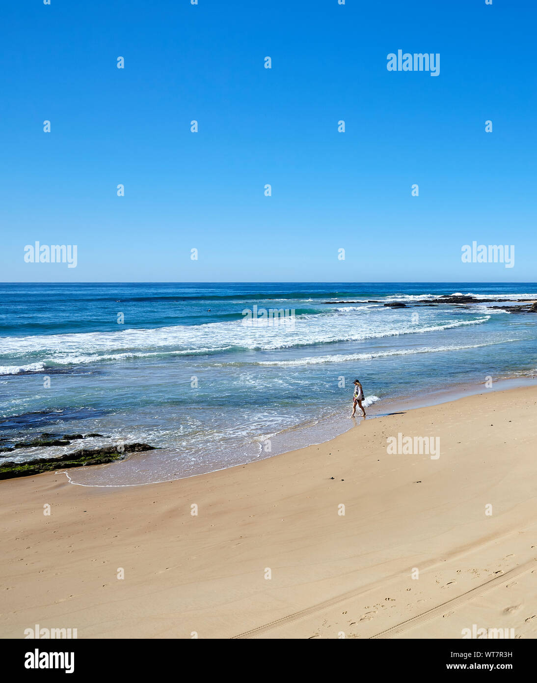Vista di una spiaggia con una persona la distanza a piedi lungo il bordo delle acque come la marea avanza su un soleggiato e limpido giorno di autunno in Australia Foto Stock