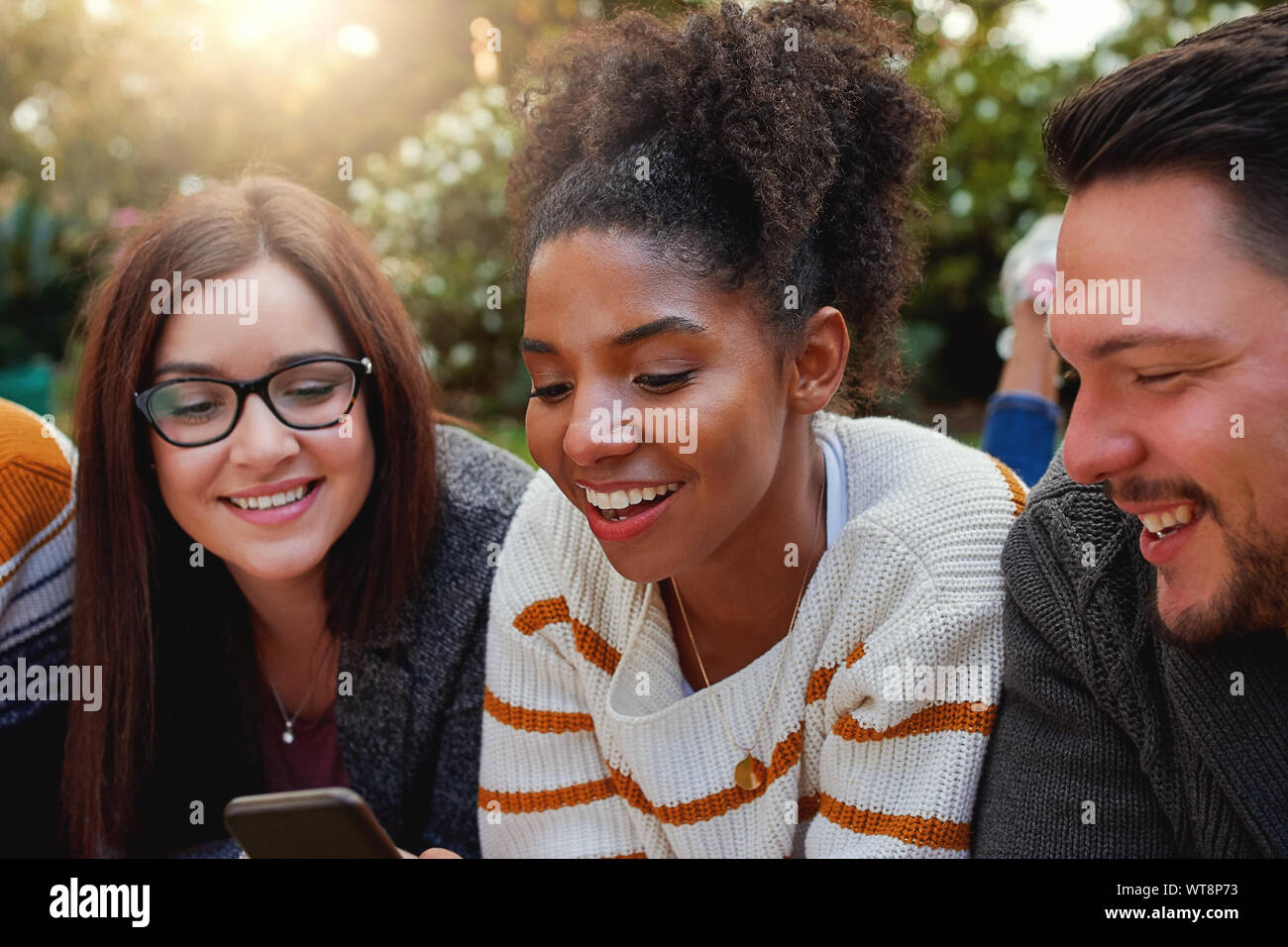Ritratto di un sorridente americano africano giovane donna con i suoi amici a guardare il telefono cellulare nel parco - caldo sole flare nella lente Foto Stock