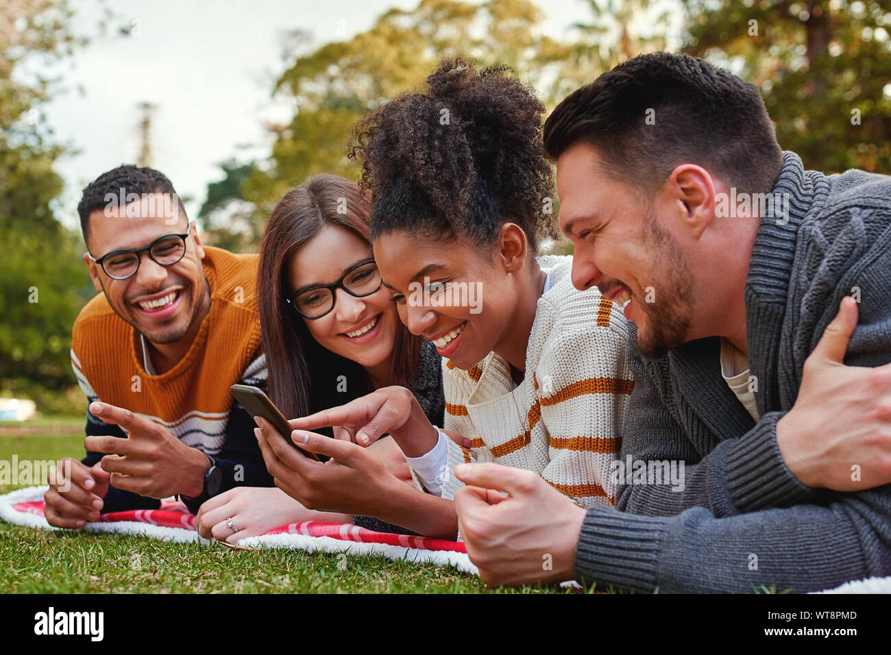 Sorridente razza mista gruppo di amici giacente insieme su erba verde utilizzando il telefono cellulare nel parco - amici avente un picnic Foto Stock