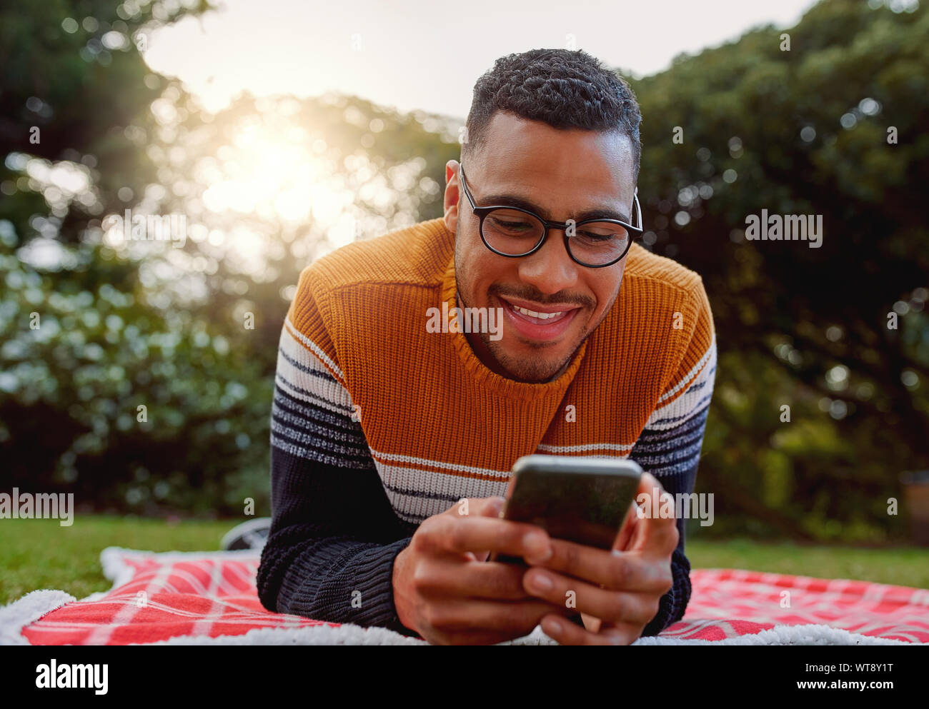 Ritratto di un africano sorridente giovane studente nero da indossare occhiali relax nel parco utilizzando smart phone - studente di college utilizzando mobile in posizione di parcheggio Foto Stock