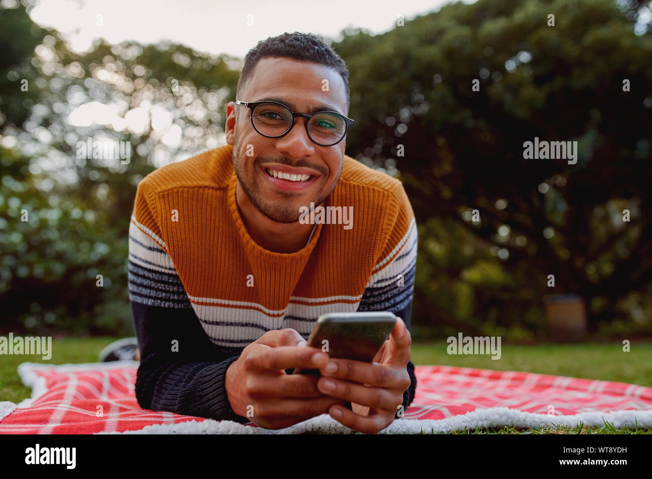 Ritratto di un sorridente americano africano giovane uomo disteso sulla coperta nel parco tenendo il telefono cellulare in mano guardando sorridente della fotocamera Foto Stock