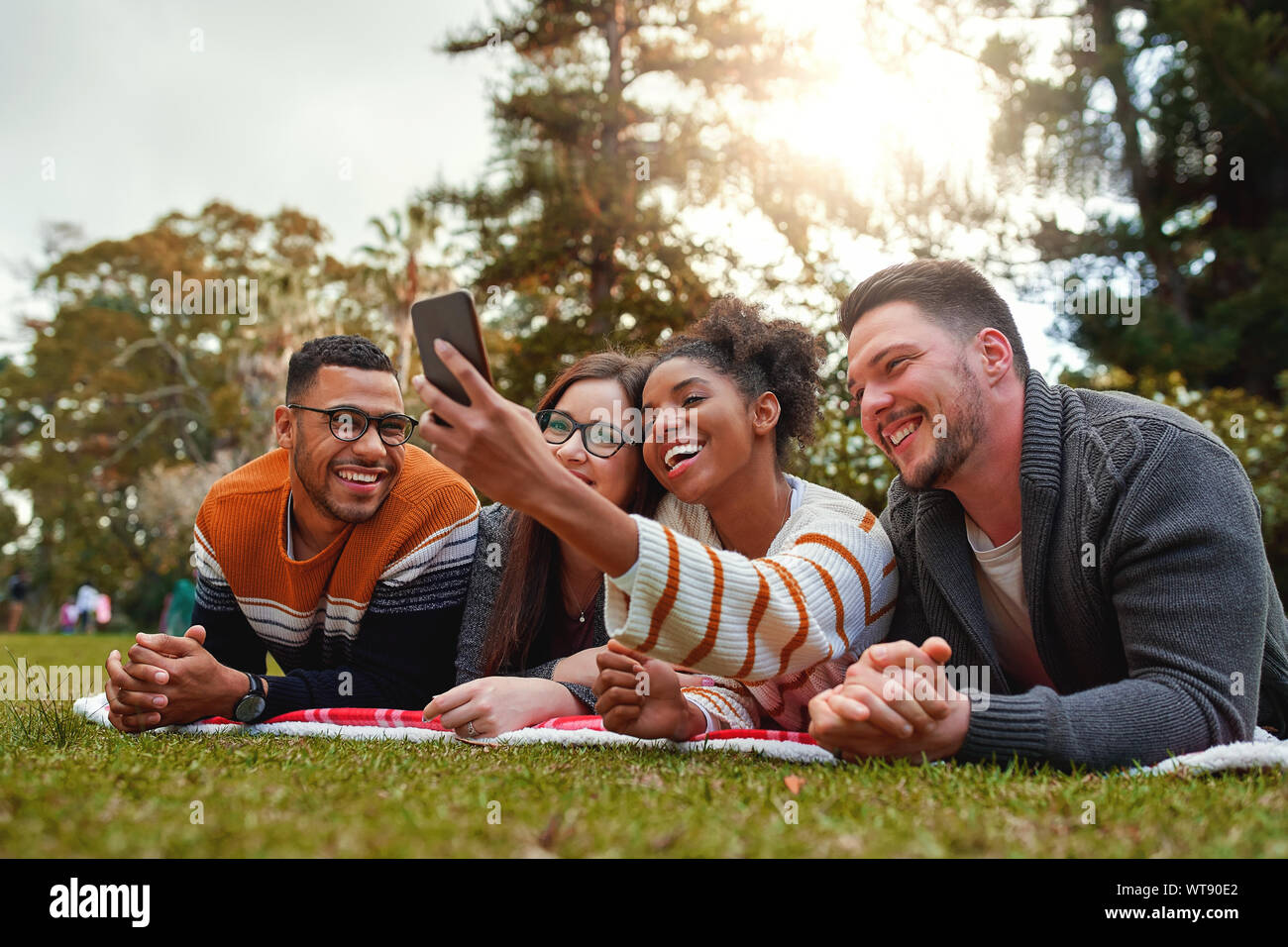 Sorridente multi etnico gruppo di amici giacente su erba verde sorridere mentre prendendo selfie sullo smartphone nel parco - molto verde della natura Foto Stock