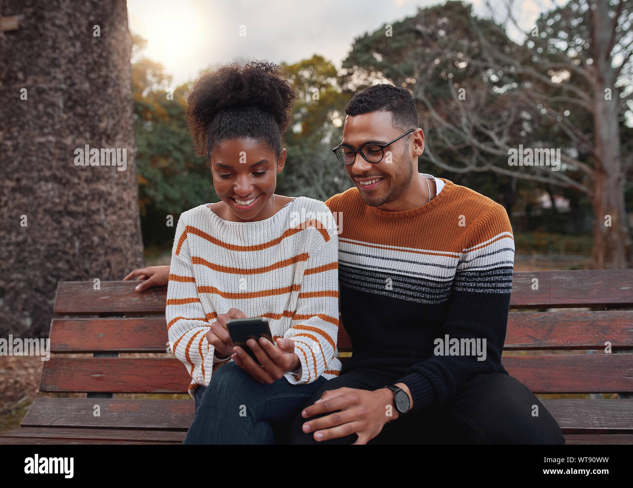 Ritratto di un africano coppia giovane siting insieme sulla panchina nel parco guardando al telefono cellulare Foto Stock