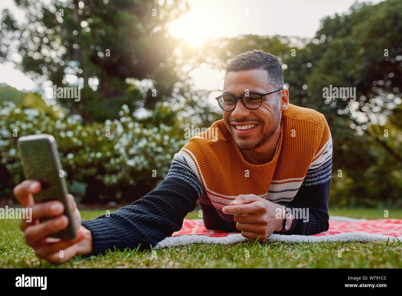 Ritratto di un giovane sorridente african american uomo disteso sulla coperta su erba verde nel parco tenendo selfie sul telefono cellulare al college - università Foto Stock