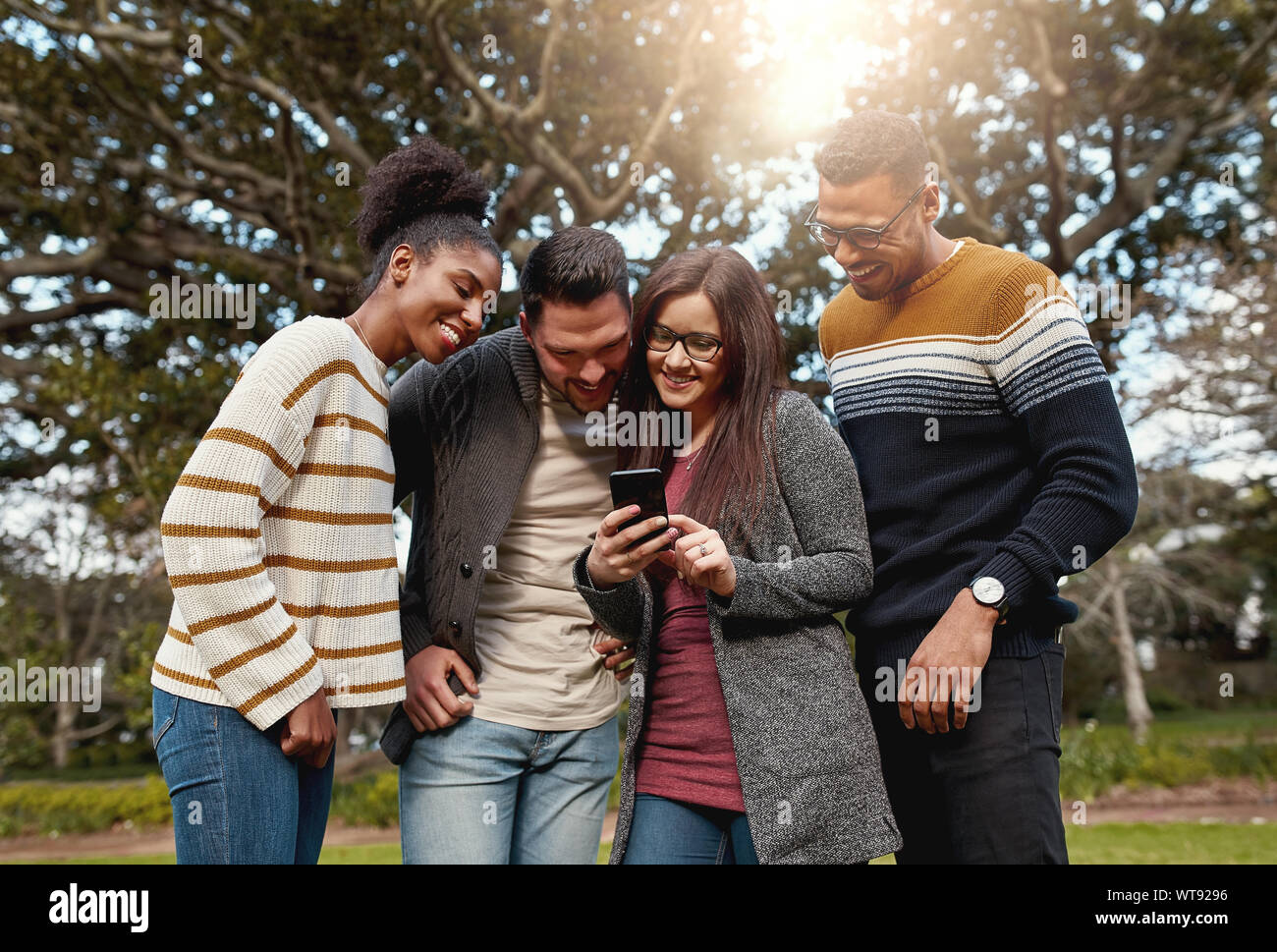 Gruppo di giovani multi razziale amici in piedi insieme guardando al telefono mobile nel parco sorridendo - caldo all'aperto Foto Stock
