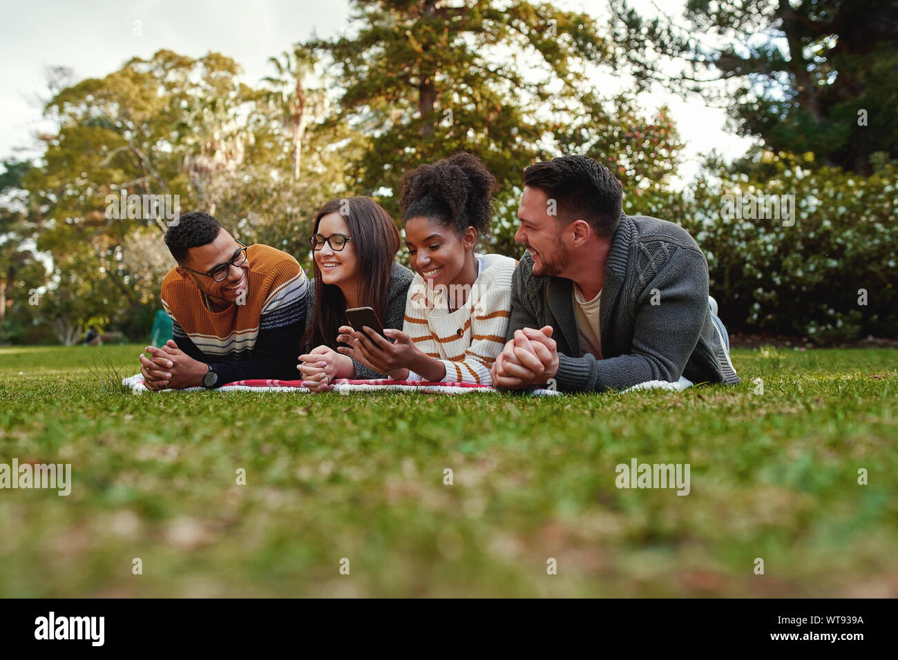 Sorridente americano africano donna giovane studente mostrando qualcosa di interessante per i suoi amici giacente insieme su una coperta sul prato verde nel parco Foto Stock