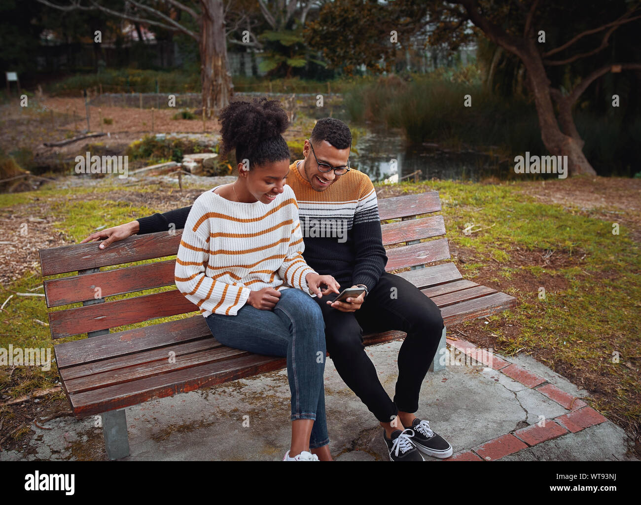 Felice giovane africano giovane seduti insieme sul banco vicino stagno nel parco utilizzando il telefono cellulare Foto Stock