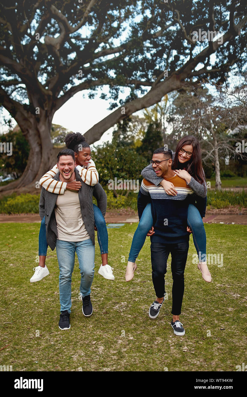 Felice giovani uomini che trasportano piggyback ride per le loro fidanzate in un parco sorridenti - divertimento all'aperto Foto Stock