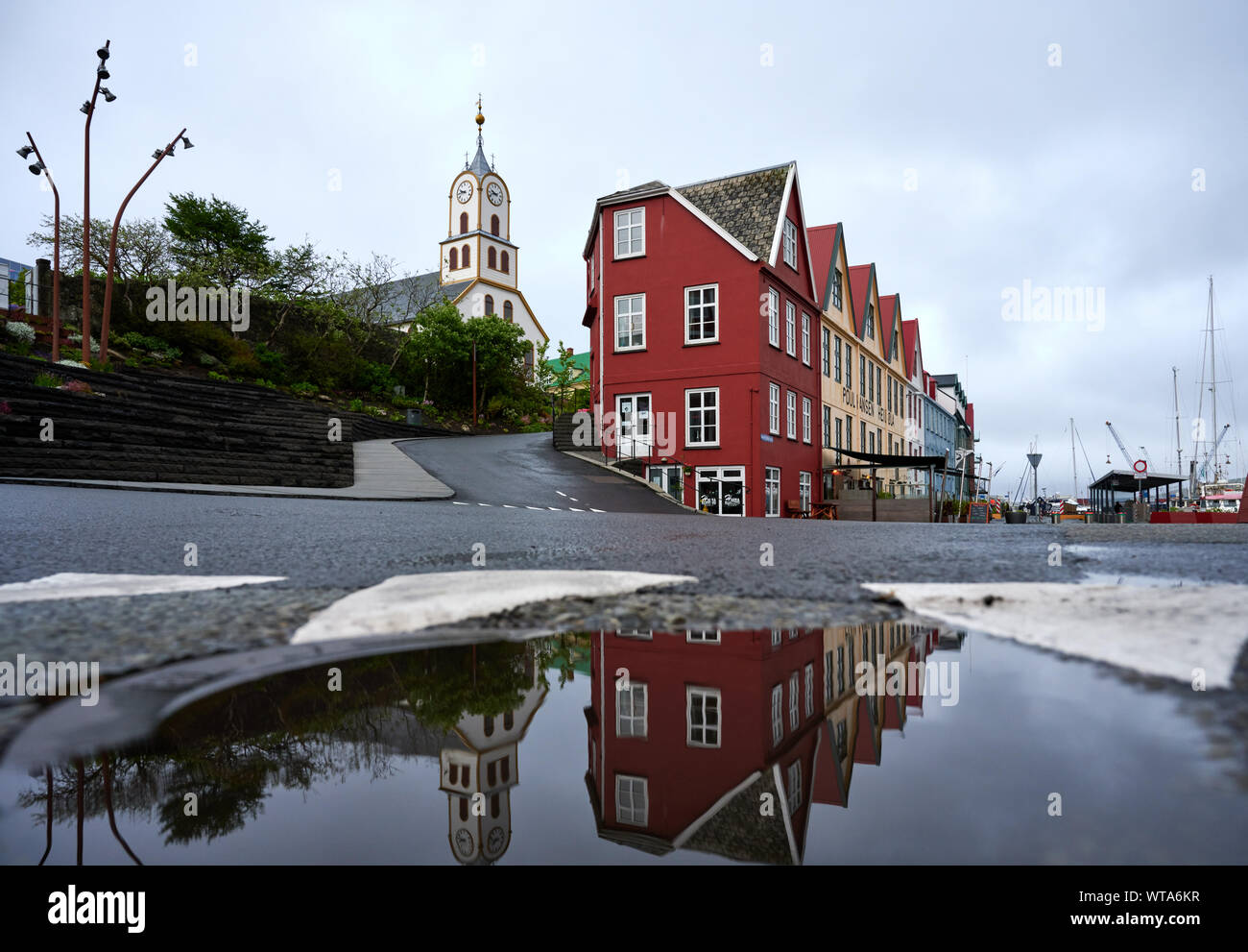 Centro storico con edifici luminoso e bianco tradizionale chiesa riflessa nell'acqua di impasto su strada sulle Isole Faerøer su giornata uggiosa Foto Stock