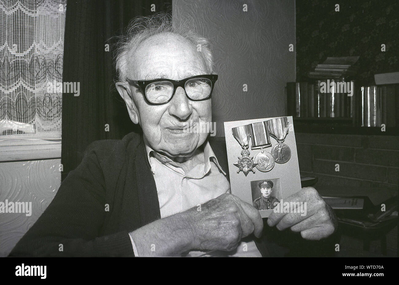 Anni ottanta, storico, un anziano British uomo seduto in una stanza, tenendo in mano una carta con i suoi tre medaglie militari e una fotografia di se stesso in uniforme come un giovane veterano britannico che ha lottato per il suo paese in WW2, Inghilterra, Regno Unito. Foto Stock