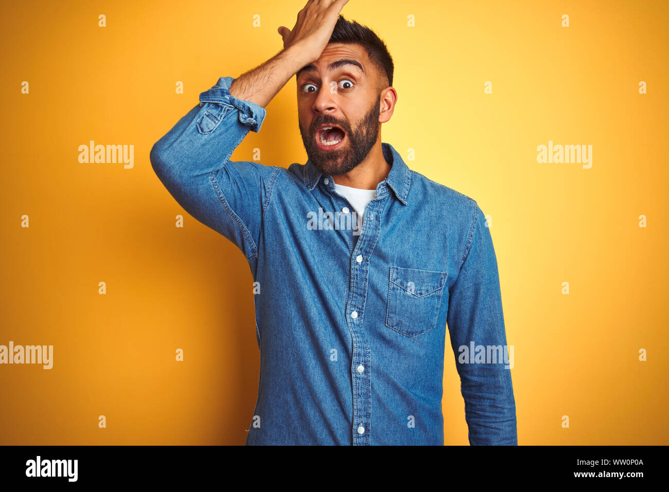 Giovani indiani uomo che indossa il denim shirt permanente isolato su sfondo giallo sorpreso con la mano sulla testa per errore, ricordati di errore. Hai dimenticato, bad mem Foto Stock