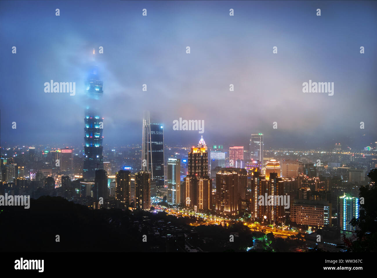 Taiwan: Misty nuvole passare la sommità del grattacielo Taipei 101 di notte illuminata da luci blu. Lo Skyline di Taipei Foto Stock